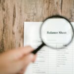 Close-up of a balance sheet document on wooden surface with a magnifying glass held by a hand.
